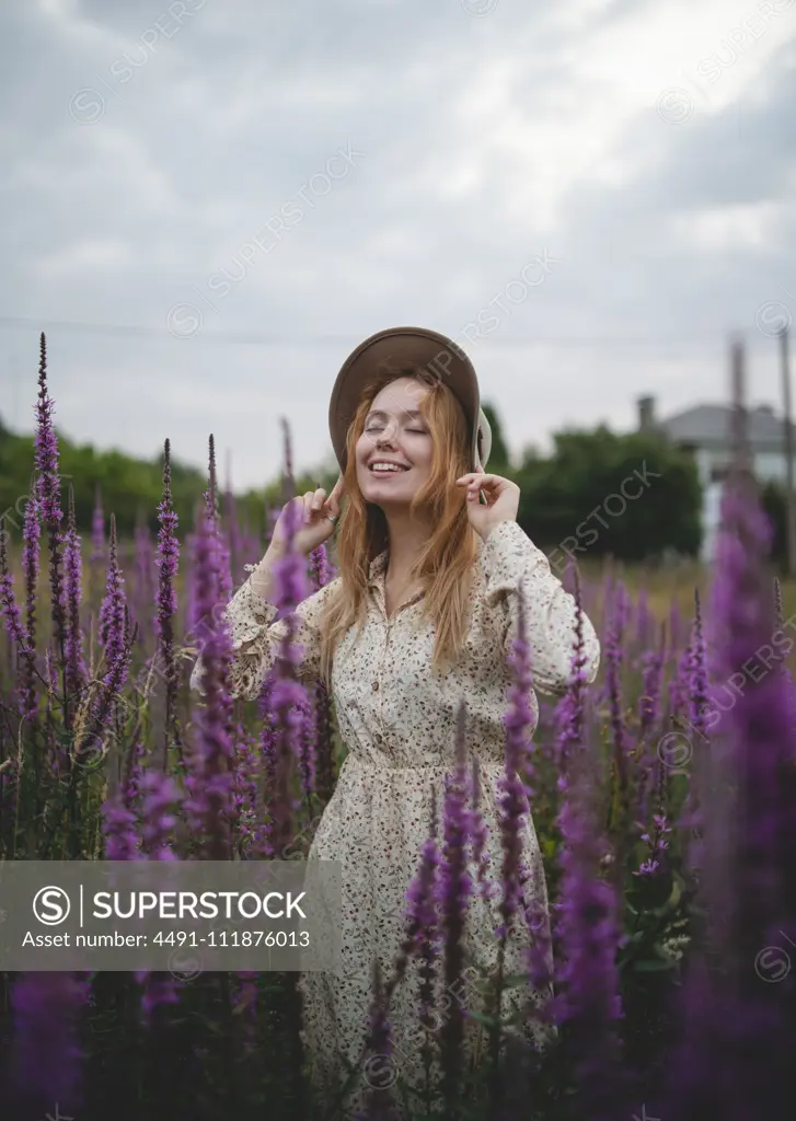 Happy sensual tender redhead lady in trendy hat with closed eyes enjoying smell of salvia blossom in meadow of Asturias, Spain;Romantic women in salvia field