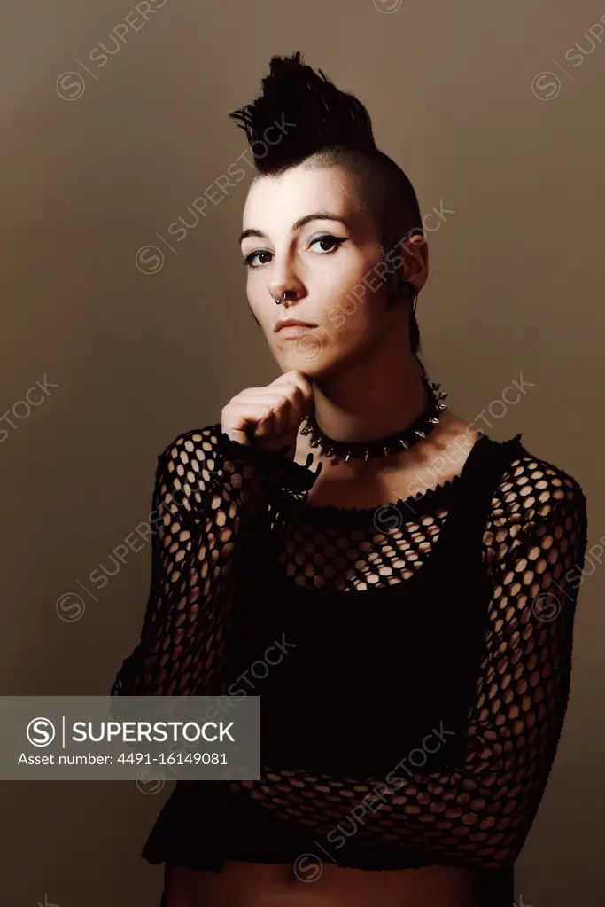 Serious adult lady with mohawk touching chin and looking at camera while standing against brown background