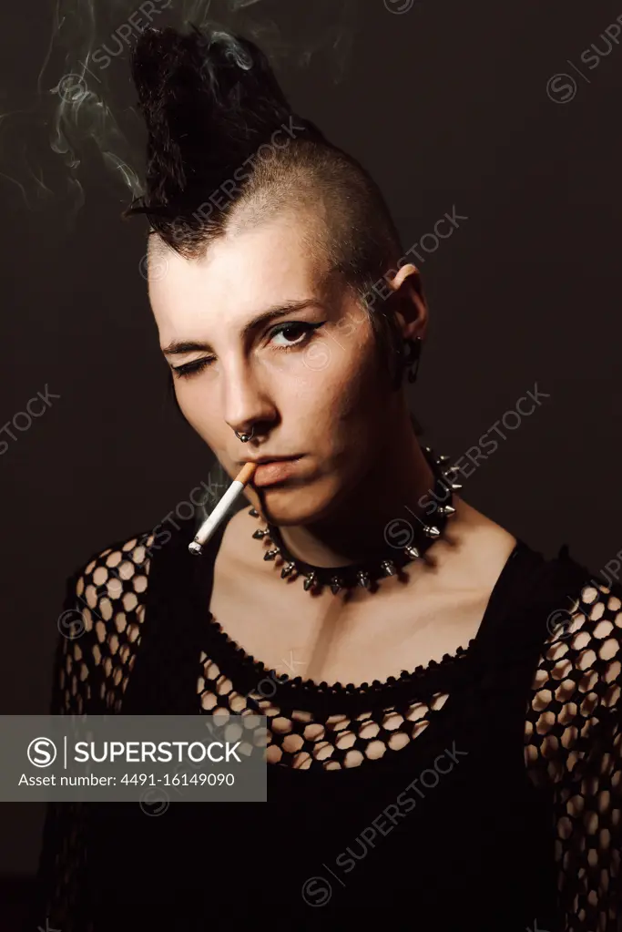 Adult female with mohawk and piercing looking at camera and igniting cigarette with lighter on black background