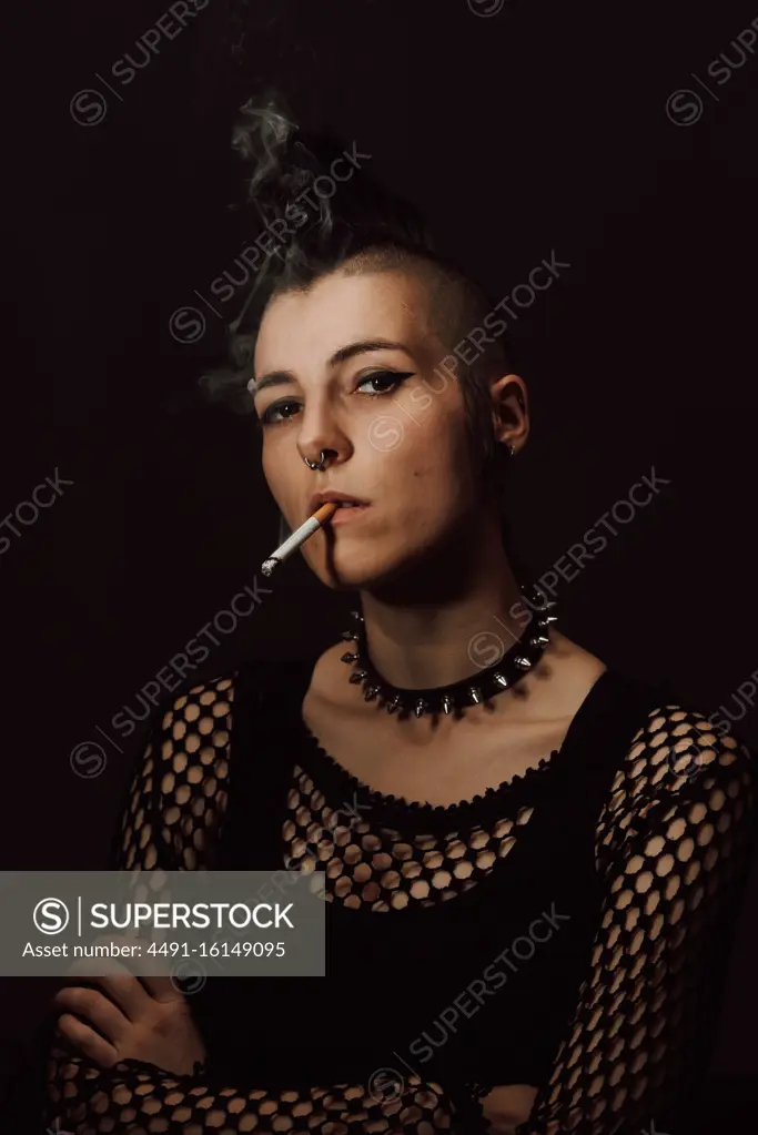 Adult female with mohawk and piercing looking at camera and igniting cigarette with lighter on black background