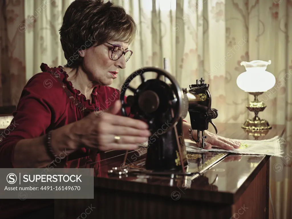 Senior lady in glasses using retro sewing machine to create linen napkin in cozy room at home