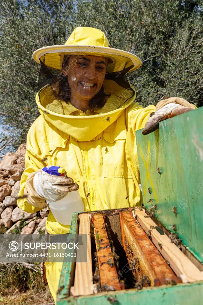 Happy female beekeeper in yellow protective wear using dispenser while spraying beehive with bees in apiary