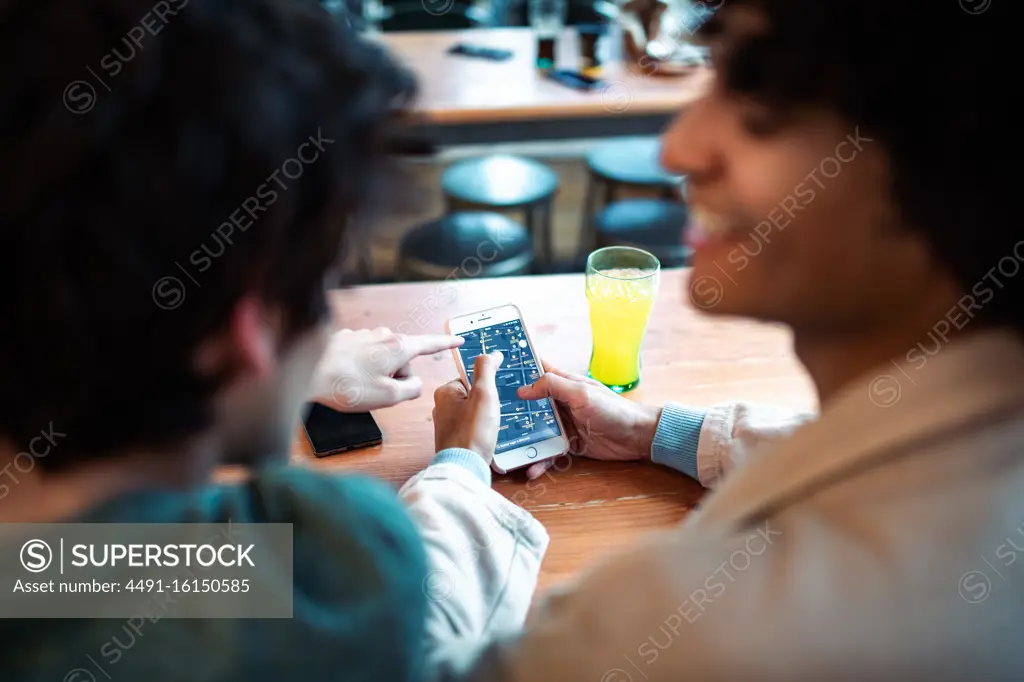 Back view of unrecognizable multiethnic young homosexual men browsing social media on smartphone and having fresh drinks smiling while sitting at cafe table during romantic date