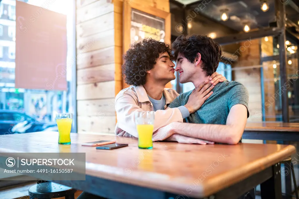 Side view of excited ethnic men embracing each other with closed eyes on table and laughing during romantic date in modern cafeteria