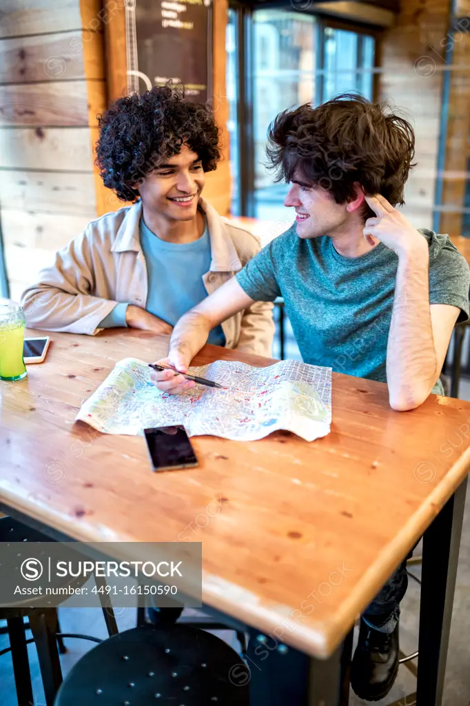 From above multiethnic young homosexual men with direction navigation map and fresh drinks smiling looking at each other while sitting at cafe table during romantic date