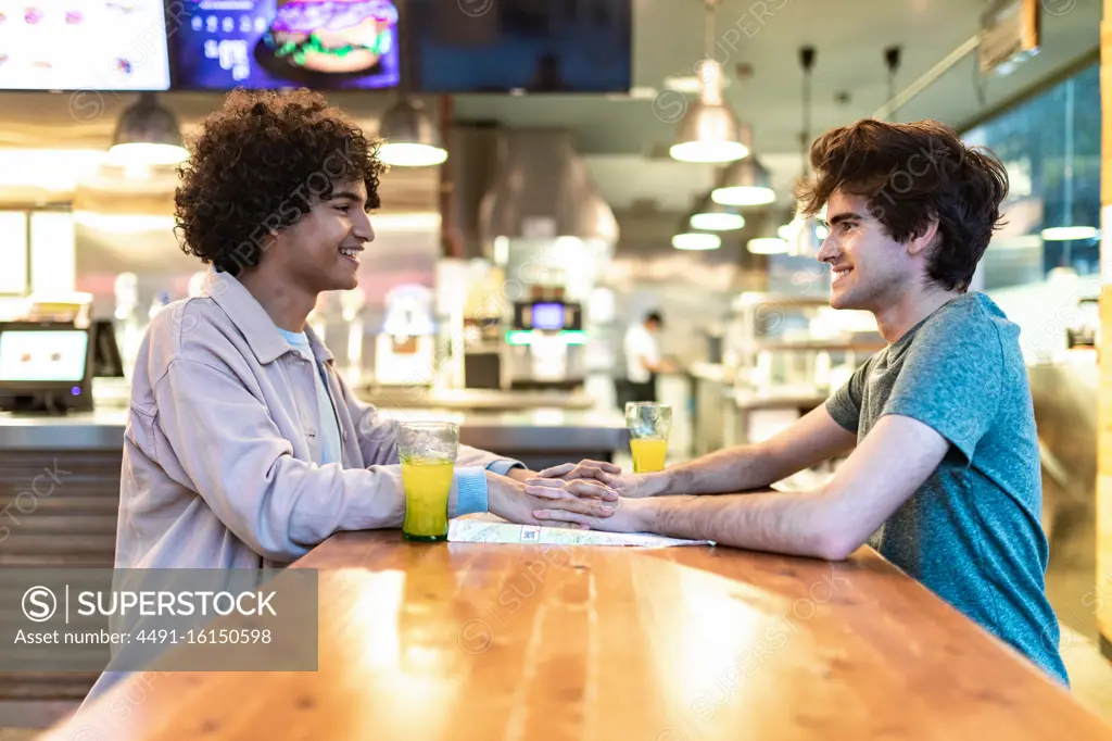 Side view of excited ethnic men embracing each other holding hands over table and laughing during romantic date in modern cafeteria