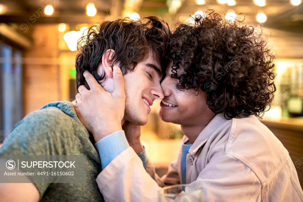 Side view of excited ethnic men embracing each other with closed eyes on table and laughing during romantic date in modern cafeteria