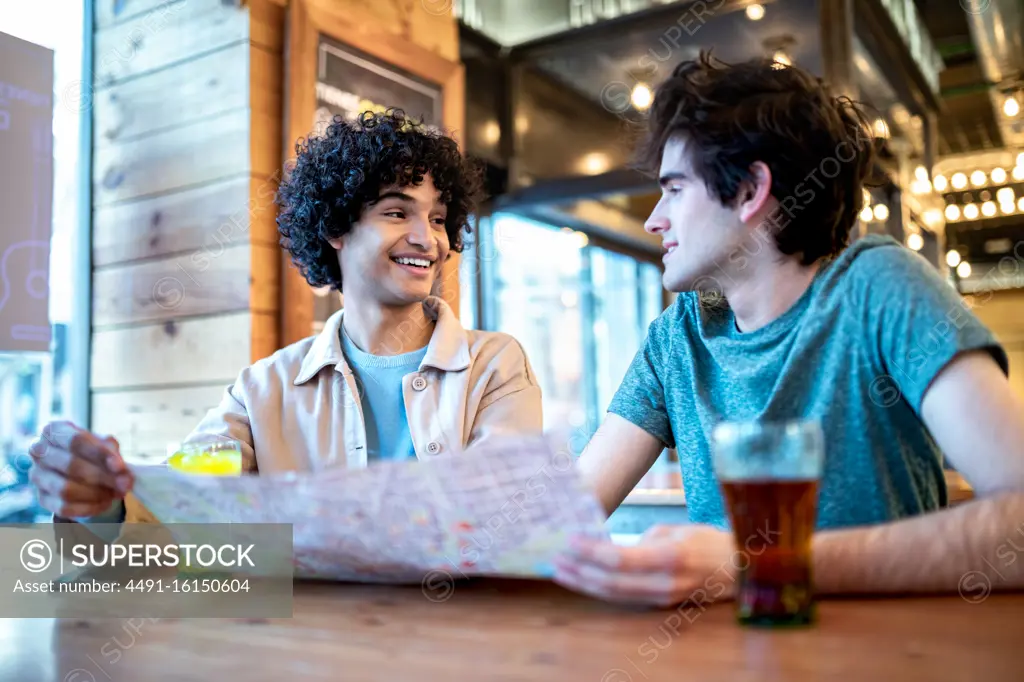 From below multiethnic young homosexual men with direction navigation map and fresh drinks smiling looking at each other while sitting at cafe table during romantic date