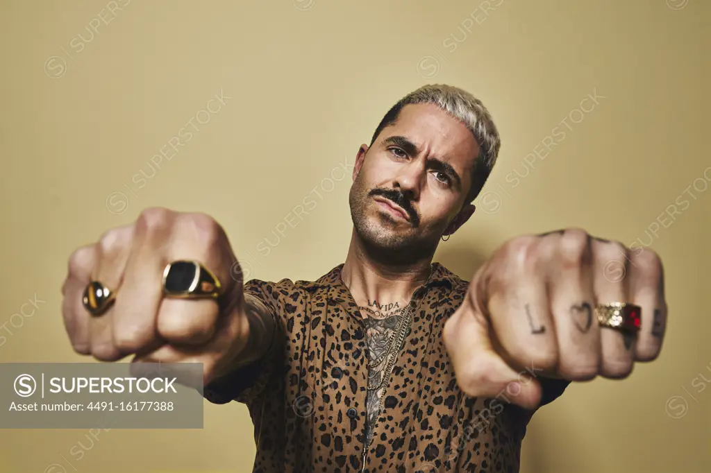 Provocative bearded guy in trendy outfit showing fists with tattoos and golden rings while standing against beige wall