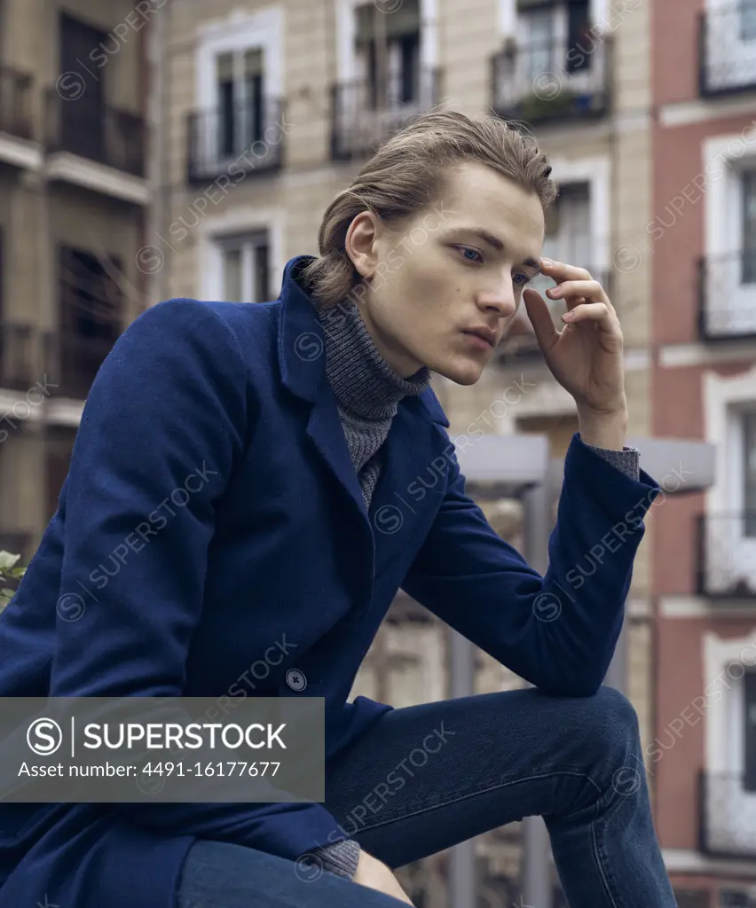 Low angle side view of serious elegant young male in stylish coat leaning on hand and thinking while sitting on street against blurred building