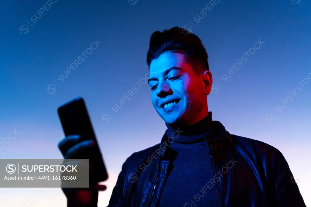 From below of happy young stylish guy in leather jacket using smartphone while standing in blue neon light against colorful sunset sky