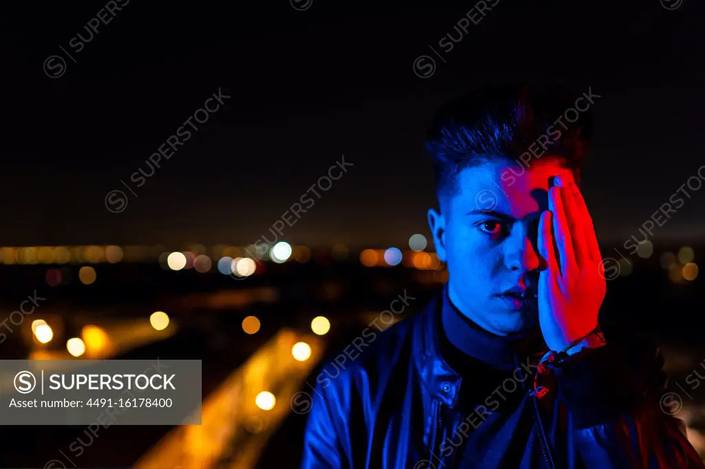 Contemporary young guy covering half of face with hand and looking at camera while standing under bright red and blue light on blurred background of city street at night