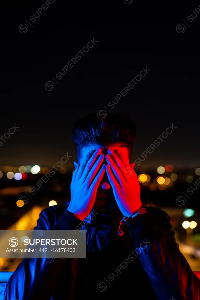 Contemporary young guy covering face with hand while standing under bright red and blue light on blurred background of city street at night