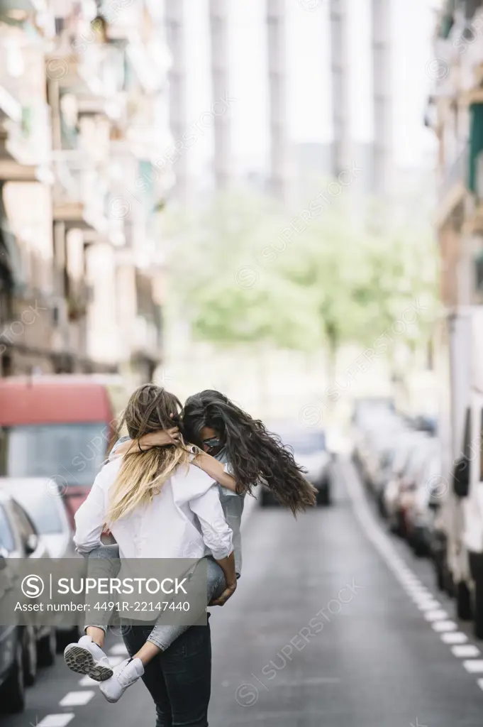 Back view of young woman carrying her girlfriend near cars on road. Vertical outdoors shot. 