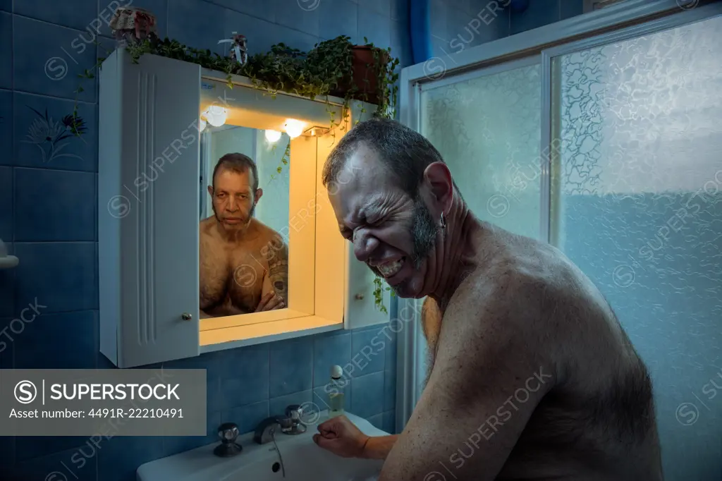 Mature man frowning from pain and stress while standing in bathroom with self reflection in mirror looking depressed