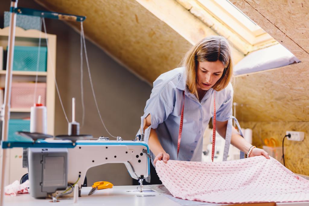 Female tailor examining pink fabric while standing near table with sewing machine in attic workshop