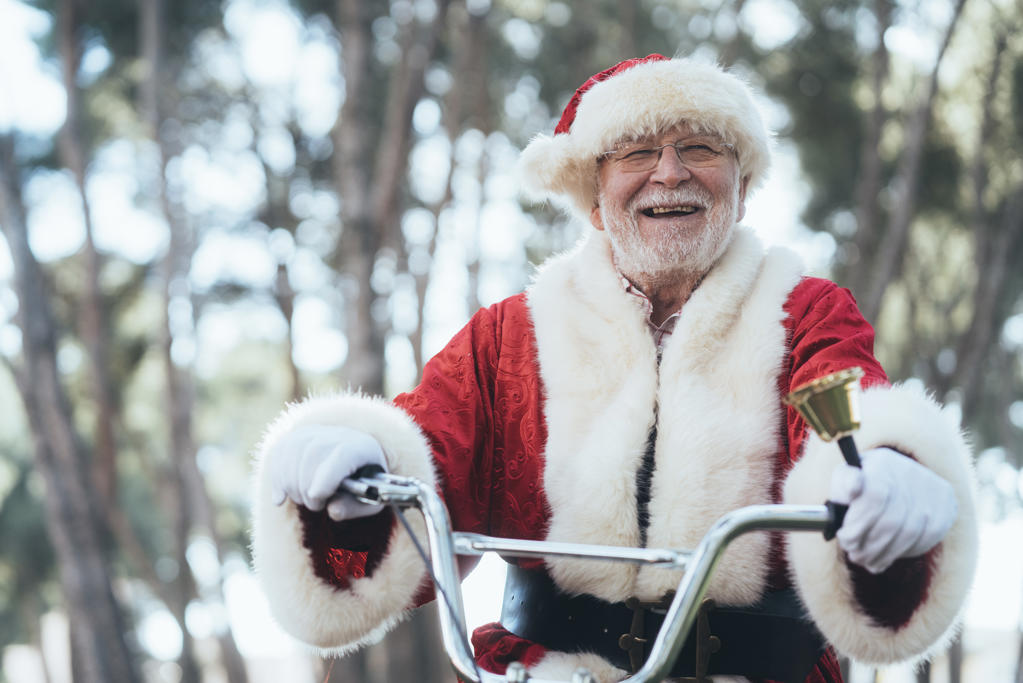 From below senior man in costume of Santa Claus sitting on cycle, ringing bell and looking at camera