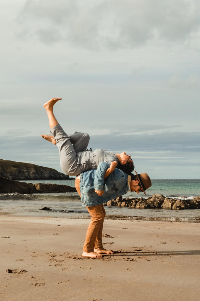 Side view of man holding on back laughing woman with raised legs while having fun together and enjoying holidays on sandy beach with rocks on cloudy day in Scotland