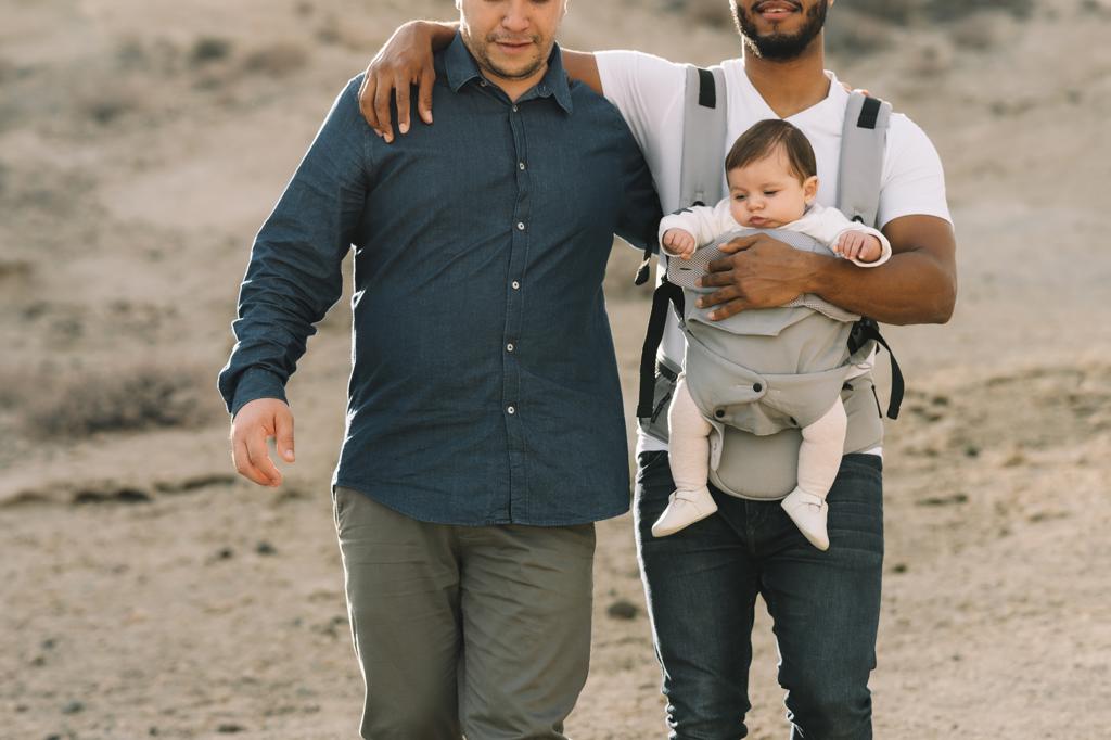 Crop black casual man holding on shoulder anonymous male boyfriend and holding little calm baby in grey carrier while strolling on nature at daytime;Unrecognizable diverse men walking with baby on nature