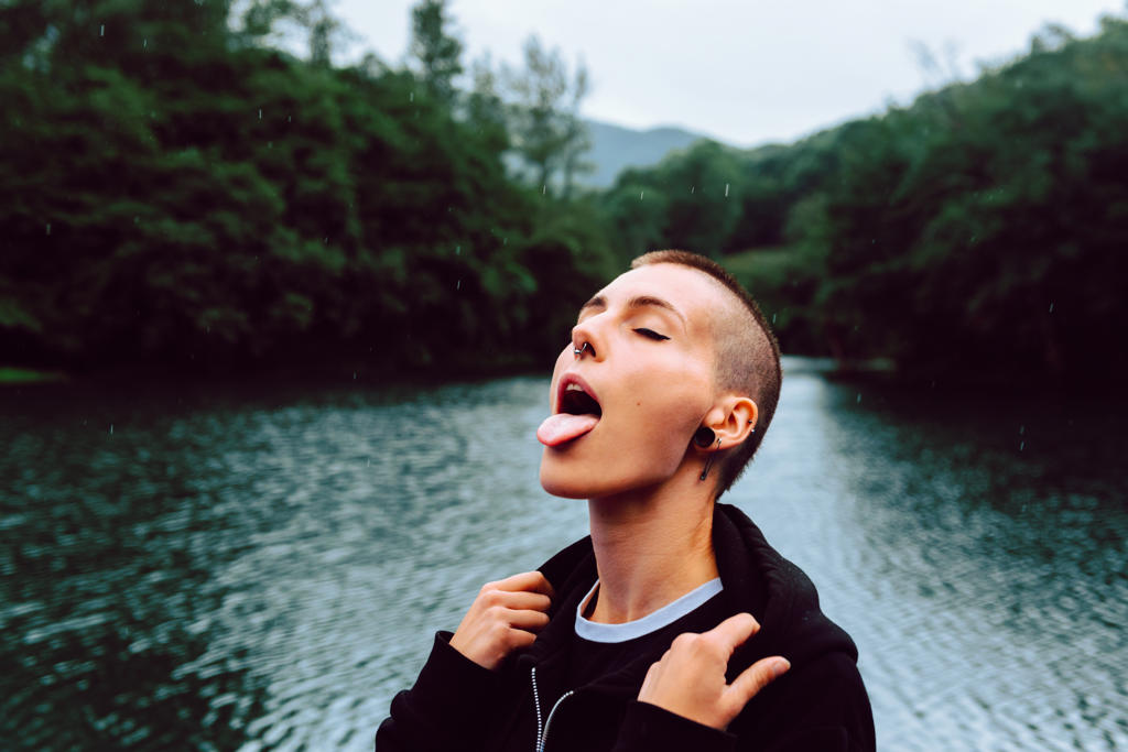 Short haired female with piercing wearing black hoodie looking up while catching raindrops with tongue near green forest and pond