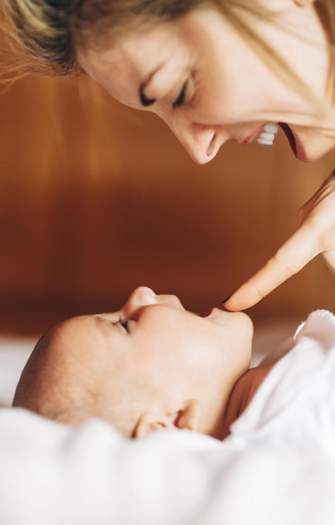 Closeup of glad mom touching nose by nose of baby playing with newborn infant with open mouth having fun lying on bed at home