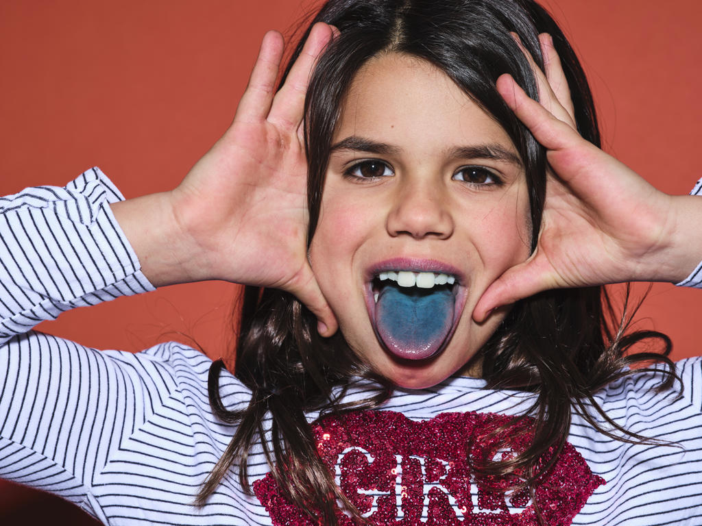 Excited preteen girl in trendy clothes looking at camera and showing blue tongue after eating colored food on red background