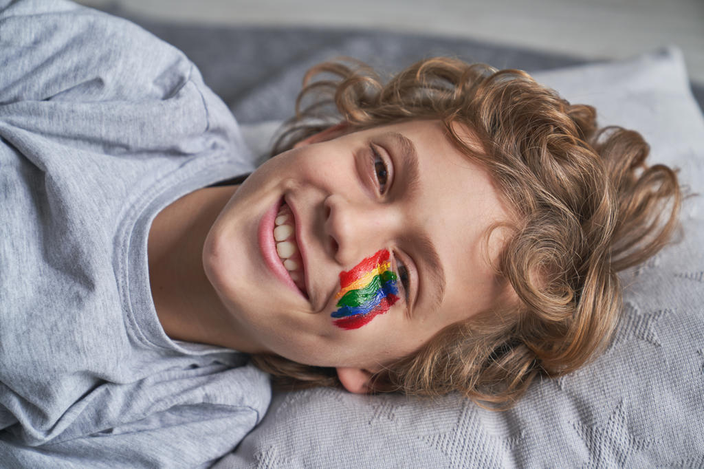 Cheerful boy with rainbow under eye smiling looking at camera while lying on blanket and pillow at home during quarantine