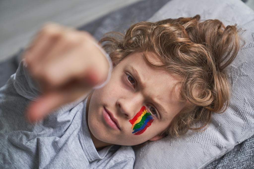 Cheerful boy with rainbow under eye smiling and pointing at camera while lying on blanket and pillow at home during quarantine
