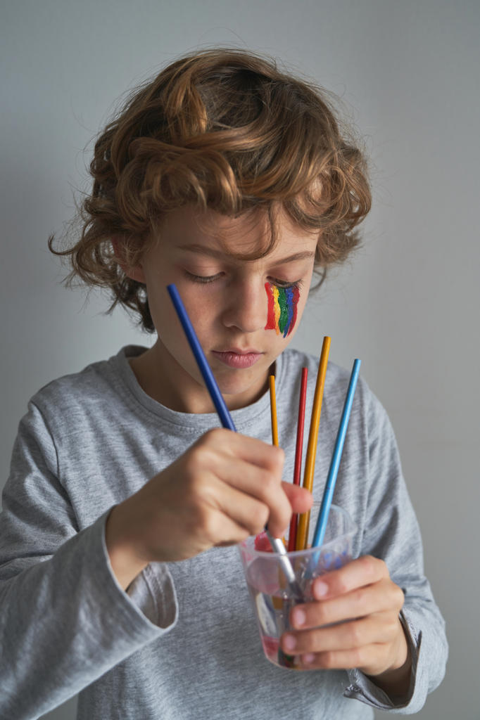 Focused boy with rainbow on face washing brush in cup of water while standing against gray background and painting during quarantine
