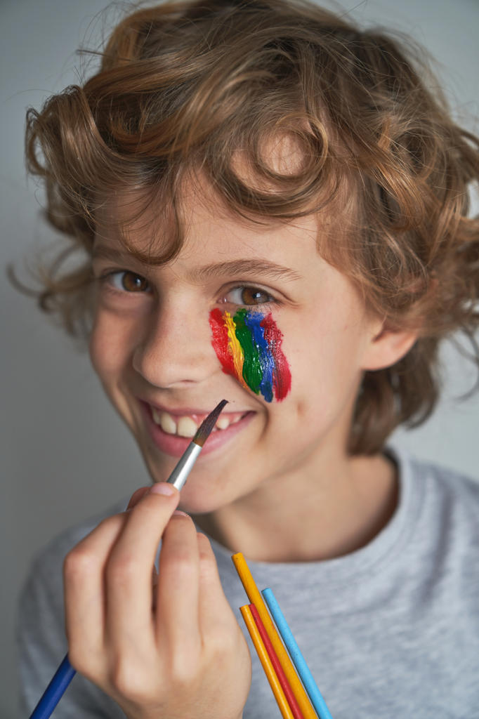 Happy boy painting colorful rainbow under eye and looking at camera while staying home during pandemic