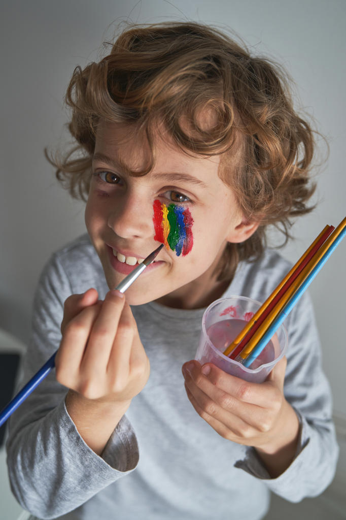 From above happy boy painting colorful rainbow under eye and looking at camera while staying home during pandemic