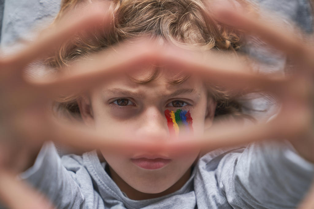 Top view of unhappy boy with colorful rainbow under eye showing stop gesture with hands with stay home inscription to the camera while lying on pillow and blanket on floor