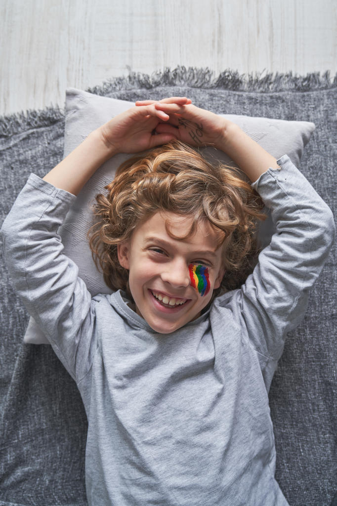 Top view of happy boy with rainbow under eye lying on blanket and pillow and laughing while staying home during quarantine