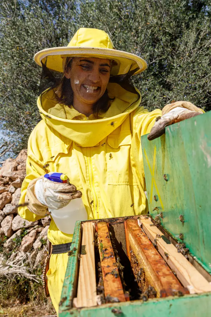 Happy female beekeeper in yellow protective wear using dispenser while spraying beehive with bees in apiary