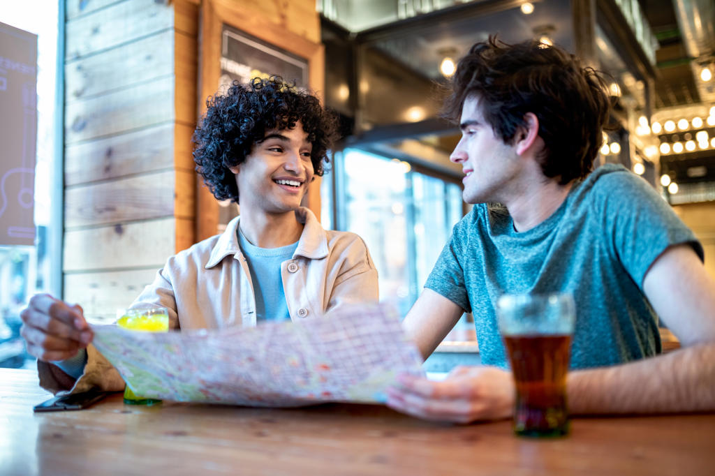 From below multiethnic young homosexual men with direction navigation map and fresh drinks smiling looking at each other while sitting at cafe table during romantic date