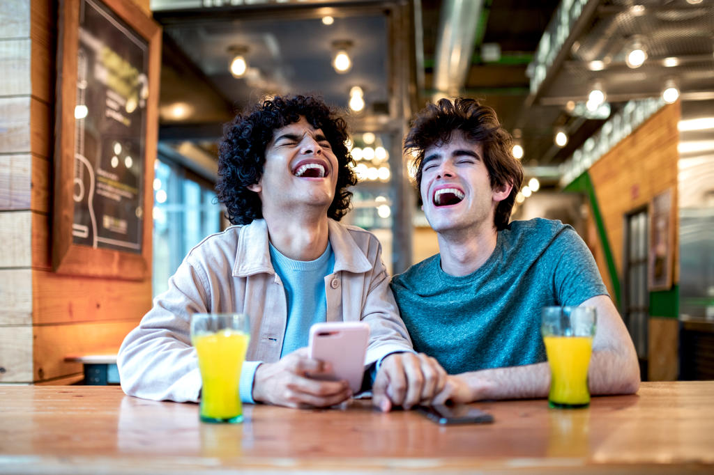 Multiethnic young homosexual men browsing social media on smartphone and having fresh drinks smiling with closed eyes while sitting at cafe table during romantic date