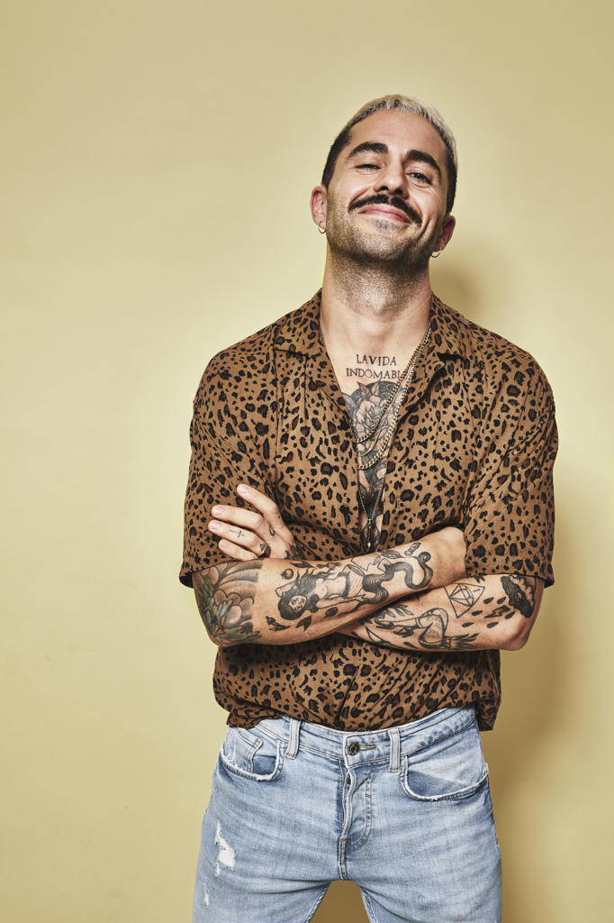Cheerful fashionable male model with tattoos wearing trendy leopard shirt and jeans standing against beige background and looking at camera