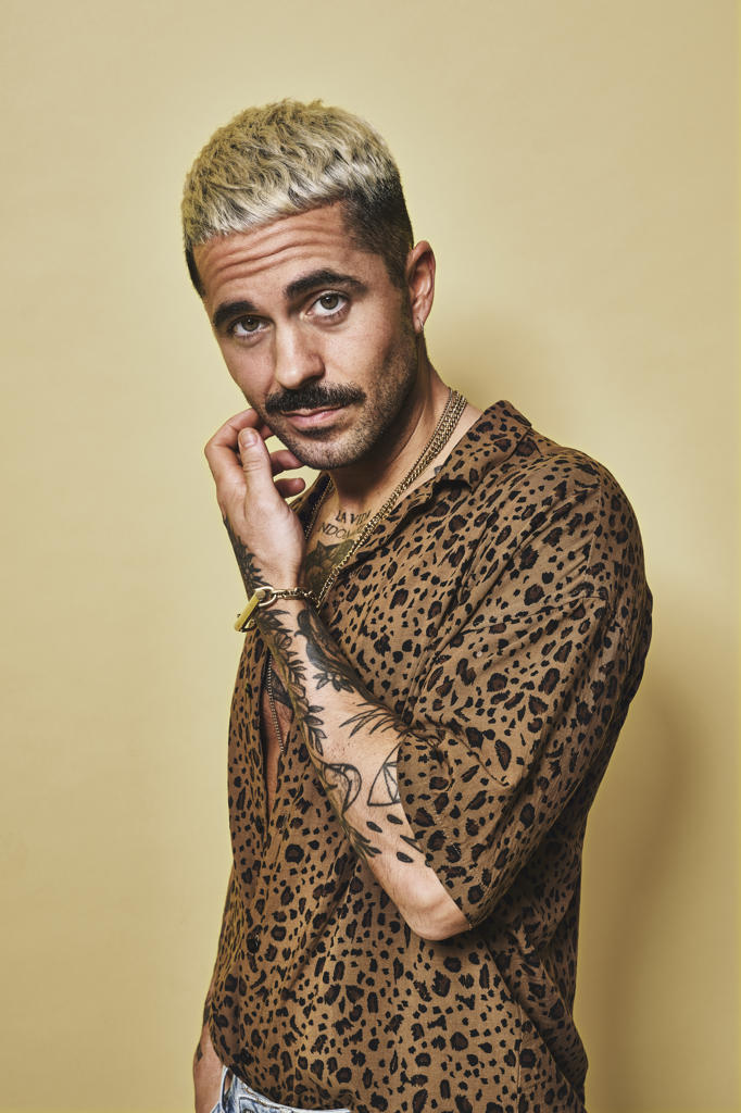 Fashionable male model with tattoos wearing trendy leopard shirt and jeans standing against beige background and looking at camera