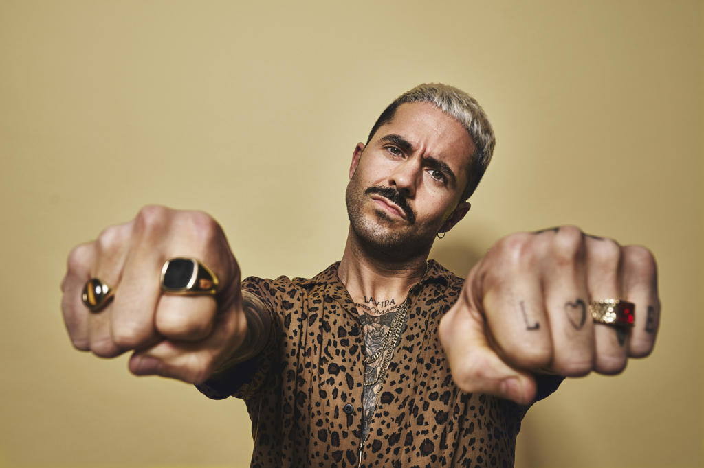 Provocative bearded guy in trendy outfit showing fists with tattoos and golden rings while standing against beige wall