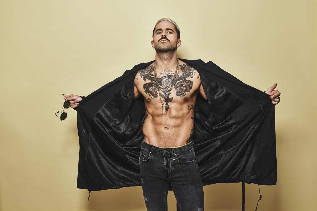Brutal muscular sexy fit male opening with hands black coat showing up naked tattooed torso while on trendy jeans with stylish sunglasses standing against beige background looking at camera