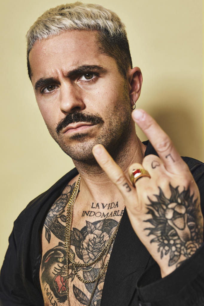 Young provocative male with mustache in black coat over naked tattooed torso showing rock sign gesture while standing against beige background looking at camera