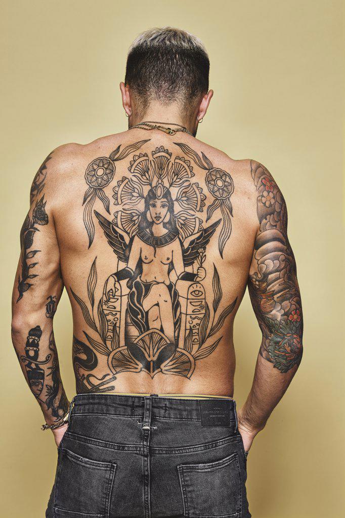 Back view of unrecognizable man with muscular tattooed body in jeans standing against beige background
