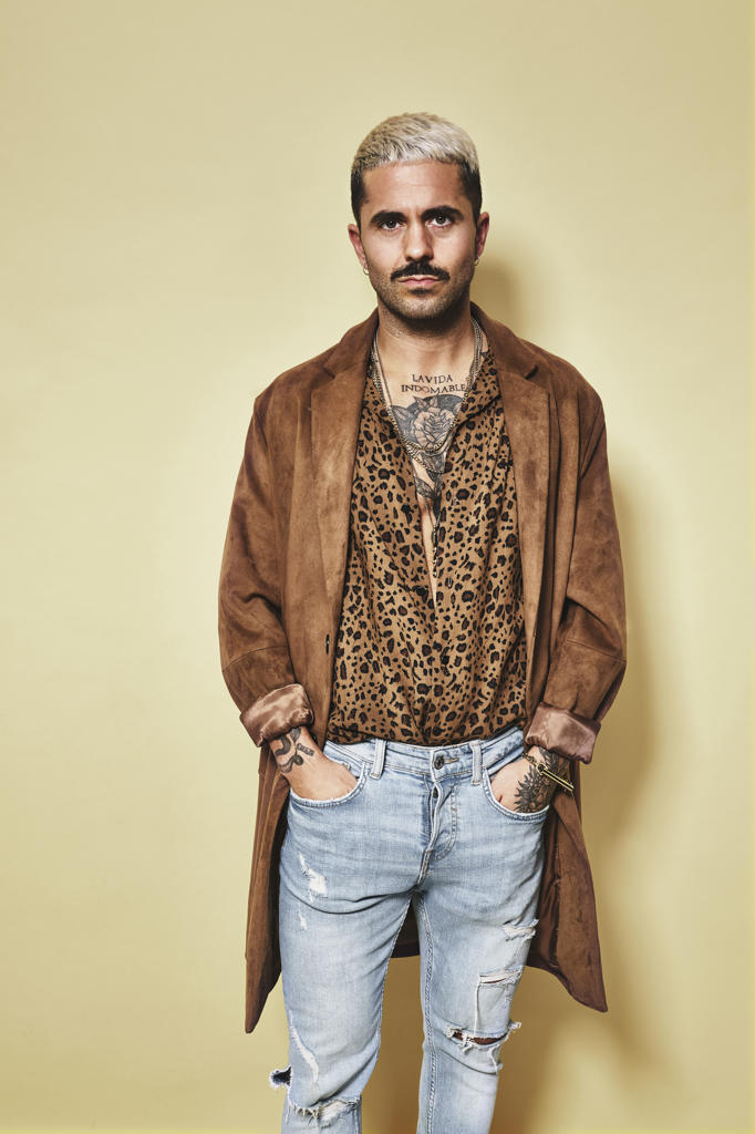 Fashionable male model with tattoos wearing trendy coat over leopard shirt and jeans standing against beige background and looking at camera