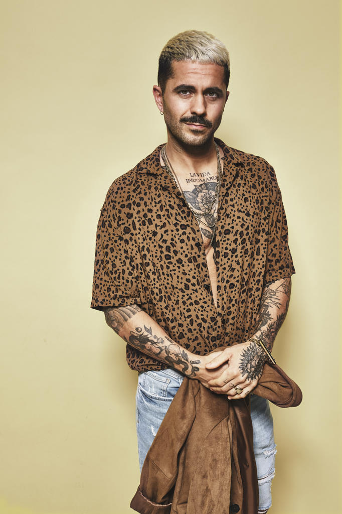 Fashionable male model with tattoos holding trendy coat over leopard shirt and jeans standing against beige background and looking at camera