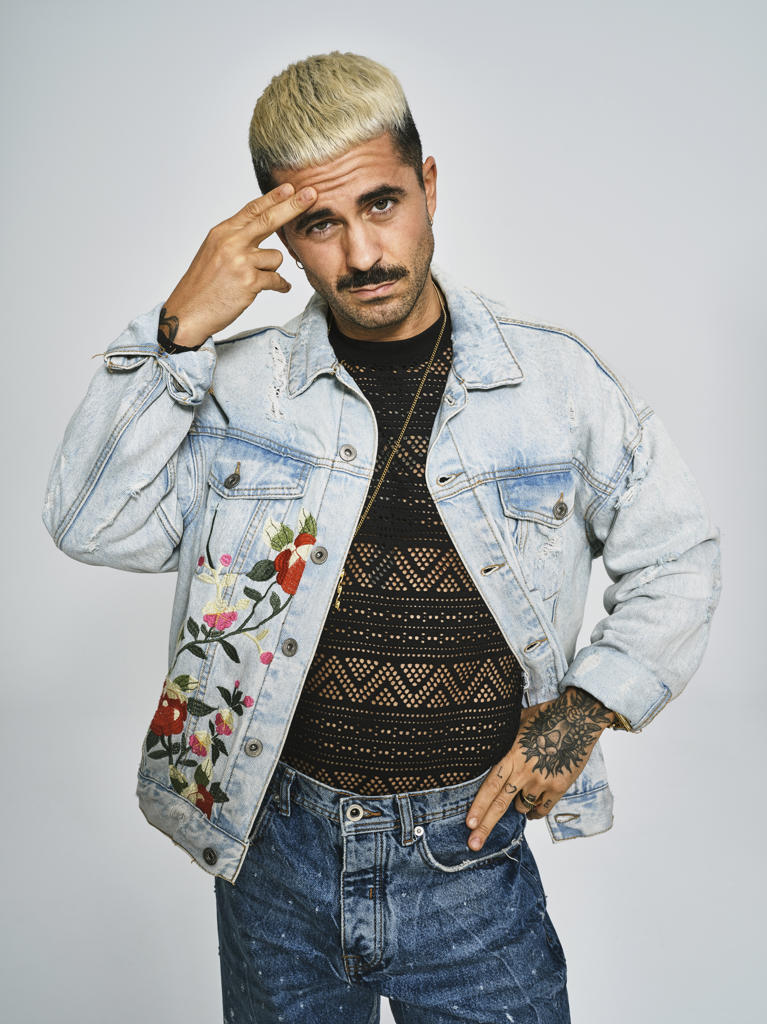 Young ethnic man making grimace doubting face with finger looking at camera wearing trendy denim jacket with floral pattern while standing against gray background