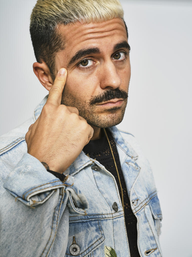 Portrait of young ethnic man making grimace doubting face with finger looking at camera wearing trendy denim jacket with floral pattern while standing against gray background