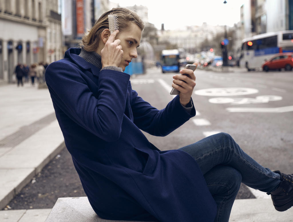 Side view of young handsome male in stylish outfit looking at smartphone and brushing hair while sitting on city street in gray autumn day