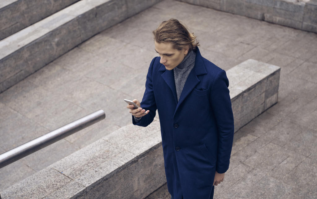 From above of serious young businessman in elegant coat checking message on mobile phone while standing on stone staircase on city street