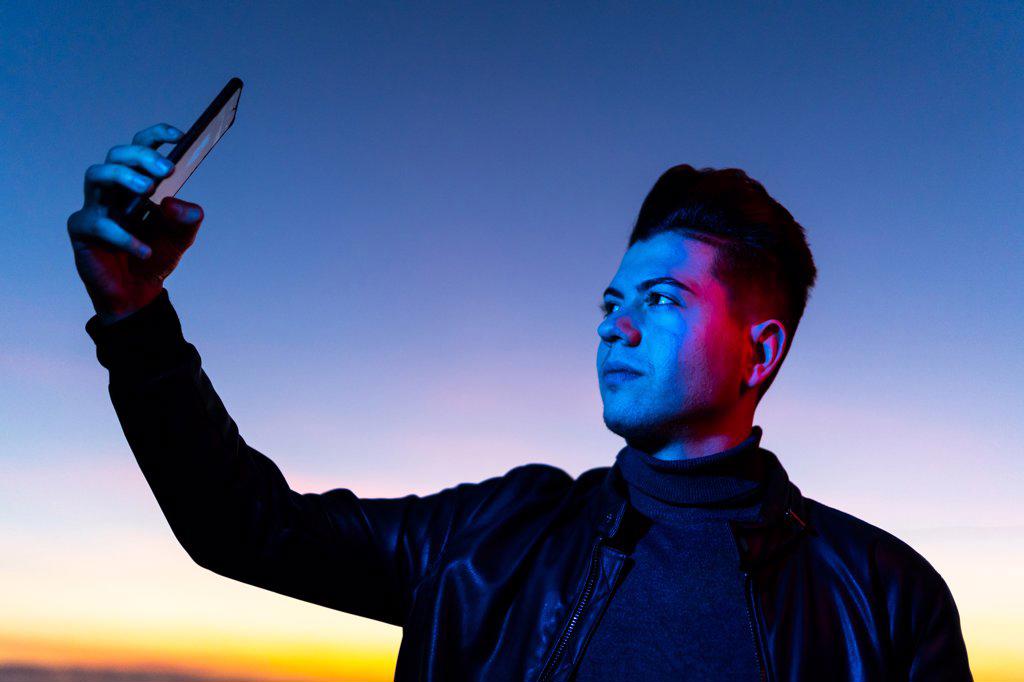 From below of serious young stylish guy in leather jacket taking selfie with smartphone while standing in blue neon light against colorful sunset sky