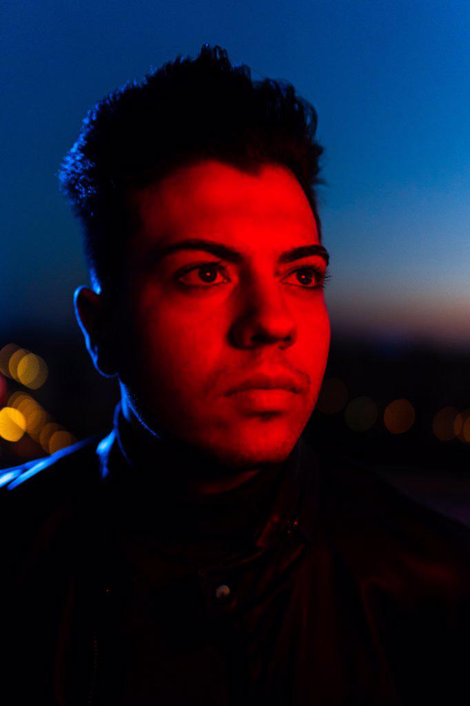 Serious young male with stylish haircut wearing black leather jacket looking away while standing on street against dark sunset sky